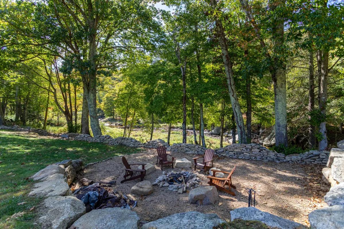 A cozy fire pit gathering spot is surrounded by Adirondack chairs and framed by a stone wall and trees. The scene feels inviting and communal, perfect for storytelling, relaxation, and enjoying nature with others.
