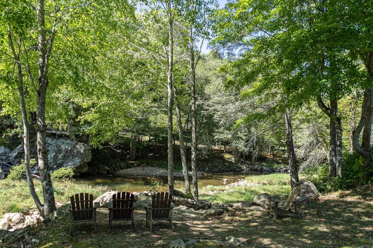 A pair of Adirondack chairs sit by a gentle stream surrounded by mature trees, offering a perfect spot to unwind. The scene captures the essence of riverside relaxation, with dappled light filtering through the leaves and a tranquil, secluded mood.