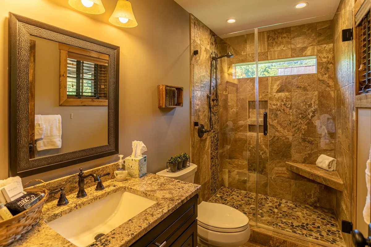 A modern bathroom featuring a granite countertop, a glass shower enclosure, a large framed mirror, and decorative lighting. The space is clean, contemporary, and calming, offering both function and style.