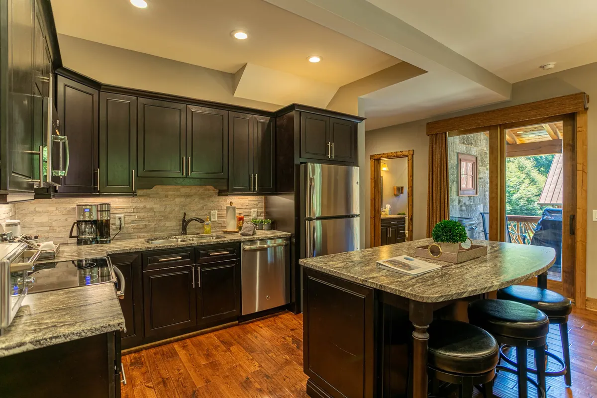 A bright, welcoming kitchen with a central island featuring three bar stools, stainless steel appliances, and natural light streaming in through the window. The open layout invites conversation and casual meals, perfectly reflecting the kitchen's role as a social hub.