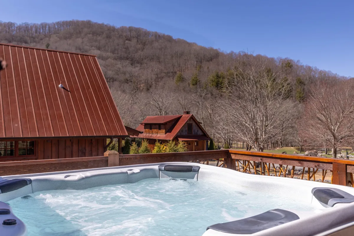 A cozy, sunlit wooden deck features a hot tub overlooking a panoramic mountain vista, with nearby cabins in view. The scene suggests moments of relaxation and connection, whether enjoying a sunset soak, stargazing, or gathering after a day of adventure.