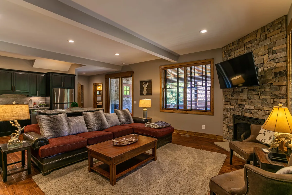 A spacious living room featuring a large sectional sofa positioned in front of a stone gas-log fireplace, accented by a wooden coffee table, a wall-mounted TV for movie nights, and a decorative lamp that adds a warm glow. The arrangement is ideal for relaxing, board games, or sharing stories by the fire.