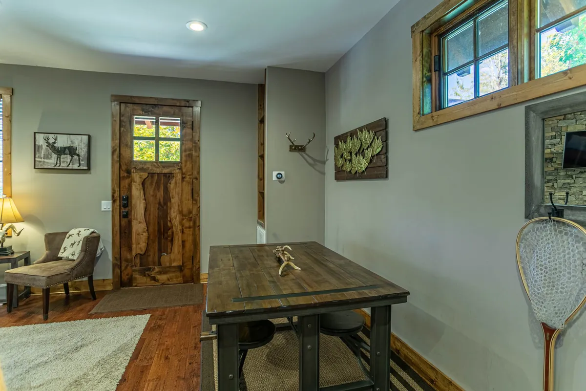 A relaxed interior scene featuring a wooden door leading to a covered patio, a dining table, antler decor, and a table lamp, evoking a warm, rustic atmosphere. The space feels connected to the outdoors, hinting at easy transitions for alfresco dining.