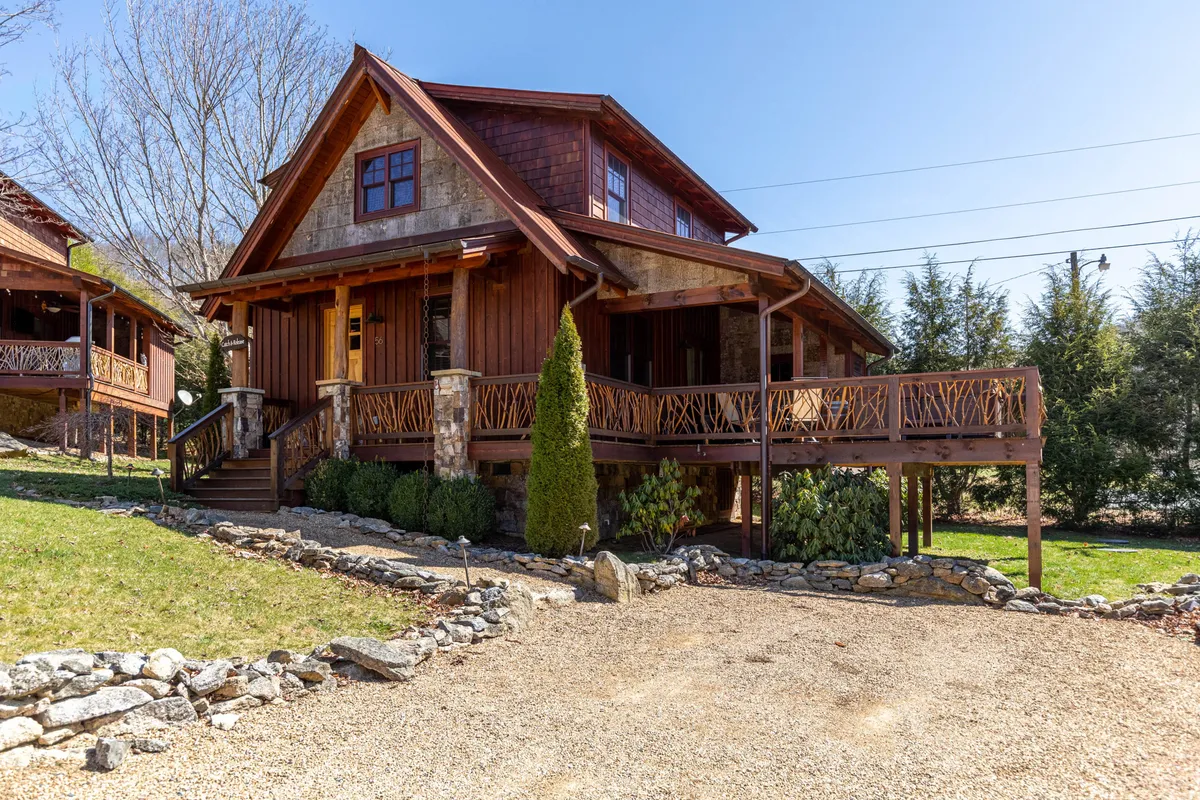 The image showcases a charming single-family cabin with a gabled roof, stone accents, and a welcoming wooden porch overlooking a beautifully landscaped garden. The porch is perfectly positioned for enjoying sunset views, and the warm lighting creates a cozy, inviting atmosphere surrounded by the natural beauty of Banner Elk.