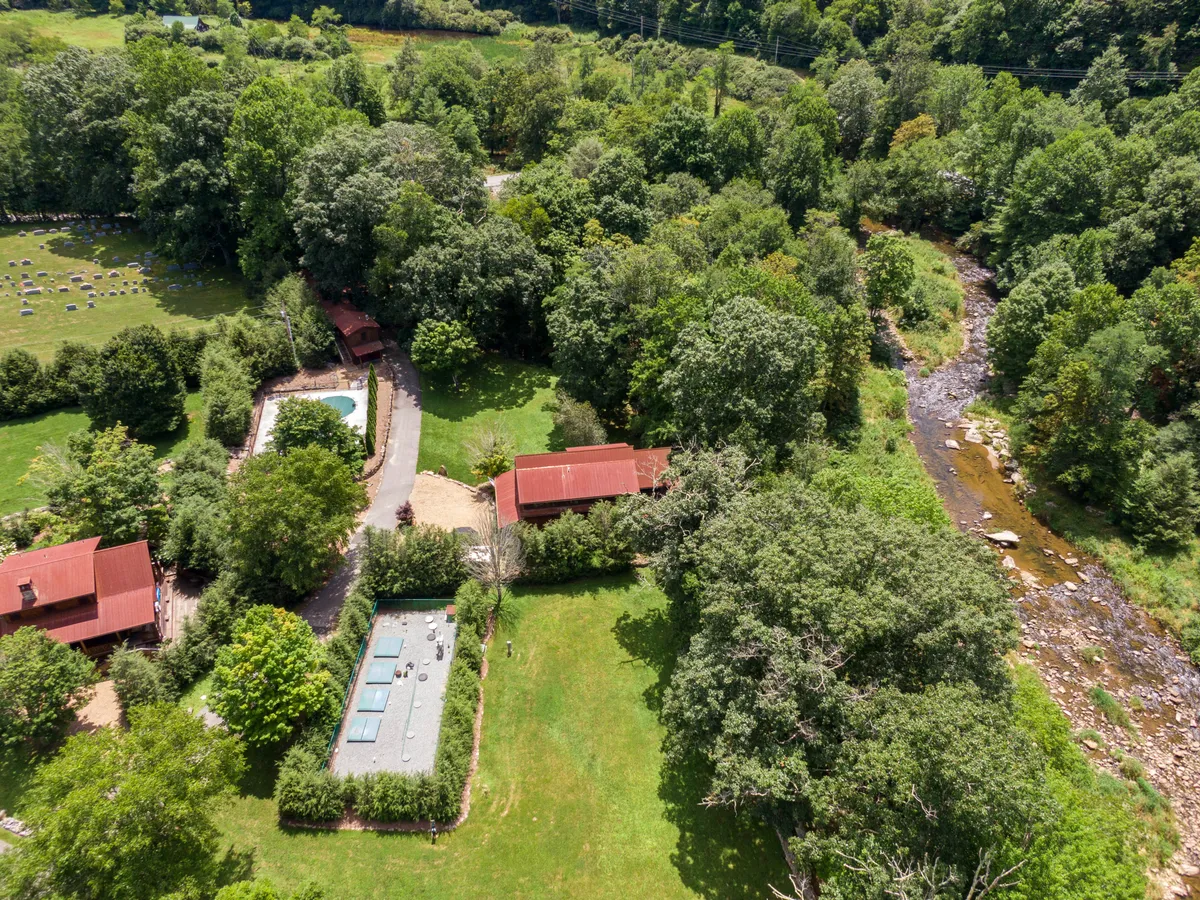 A wide exterior shot featuring houses with red roofs, a swimming pool, a gently flowing river, and abundant greenery. The scene evokes leisure, exploration, and the beauty of the cabin's riverside location.